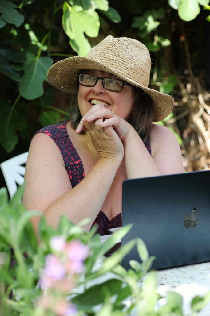 Photograph of author and artist Emily Tellwright wearing a straw hat and sitting with a laptop outside with foliage behind.